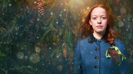 Anne Shirley, personagem da série Anne with an E, aparece com cabelo ruivo e vestido azul em meio a árvores e pinhas, em cenário natural iluminado.
