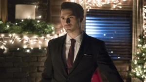 Enzo St. John aparece usando terno preto e gravata vinho em frente a uma casa decorada com luzes de Natal em cena de The Vampire Diaries.