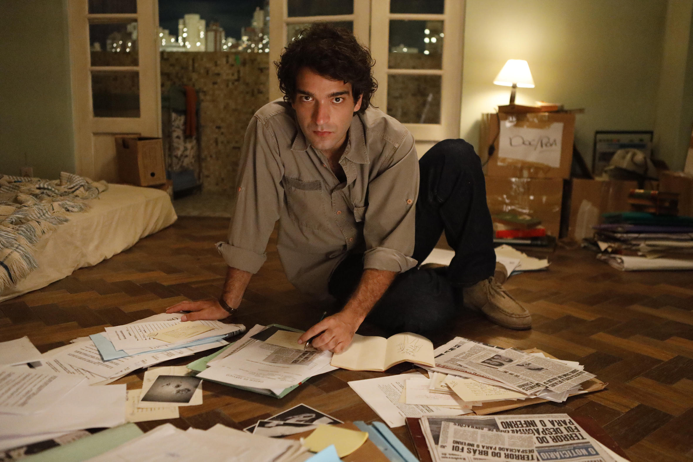 O ator Humberto Carrão aparece sentado no chão entre documentos e jornais como Caco Barcelos em cena de Rota 66 - A Polícia que Mata