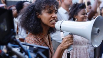 A atriz Naruna Costa é vista como Anabela falando em um megafone durante um protesto em cena de Rota 66 - A Polícia que Mata