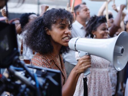 A atriz Naruna Costa é vista como Anabela falando em um megafone durante um protesto em cena de Rota 66 - A Polícia que Mata