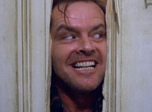 Filmes de terror psicológico: sugestões intensas para quem ama o gênero Cena icônica de O Iluminado em que Jack Nicholson, com expressão insana e sorriso ameaçador, espreita através de uma porta quebrada.