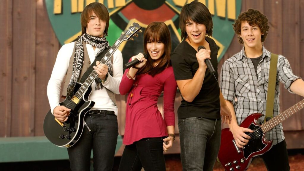 Camp Rock 3 vem aí! Joe Jonas anuncia a produção do terceiro filme Cena promocional do filme Camp Rock, mostrando quatro personagens principais em frente a um cenário de palco. Dois deles seguram guitarras, enquanto os outros dois cantam sorridentes para o público.
