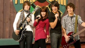 Cena promocional do filme Camp Rock, mostrando quatro personagens principais em frente a um cenário de palco. Dois deles seguram guitarras, enquanto os outros dois cantam sorridentes para o público.