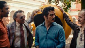 Cena do filme O Agente Secreto: quatro personagens conversam em frente a um orelhão amarelo em uma rua ensolarada. O homem ao centro veste camisa azul e bigode marcante, cercado por outros três homens em trajes típicos dos anos 70, reforçando o clima de suspense e espionagem da obra.
