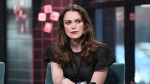 Keira Knightley, mulher de cabelos castanhos longos e soltos, usando blusa preta translúcida, está sentada em ambiente interno com fundo desfocado e iluminação suave, olhando para o lado com expressão séria.