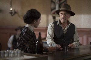 Cena do filme Dead for a Dollar (2022) mostra Christoph Waltz como Max Borlund, sentado em um bar e conversando com Rachel Brosnahan, que interpreta Rachel Price. Ele veste colete escuro e chapéu marrom, enquanto segura um copo de bebida sobre o balcão de madeira. O ambiente tem iluminação suave e decoração típica de um saloon do século XIX, evocando a atmosfera clássica dos faroestes.