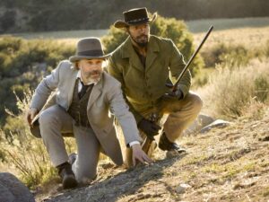 Cena ao ar livre com Christoph Waltz como Dr. King Schultz e Jamie Foxx como Django. Ambos estão agachados em uma colina, observando algo à frente. Schultz usa terno cinza claro, colete e chapéu-coco, enquanto Django veste casaco verde-oliva e chapéu de cowboy preto, segurando um rifle. A paisagem ao fundo mostra vegetação rasteira e árvores iluminadas pelo sol.