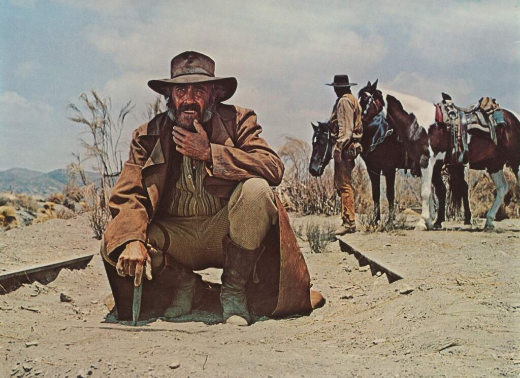 Charles Bronson como Harmonica em Era uma Vez no Oeste (1968), ajoelhado sobre a plataforma da estação ferroviária no deserto, vestindo sobretudo escuro e chapéu de aba larga, tocando sua gaita enquanto encara os pistoleiros sob o sol intenso do Velho Oeste.