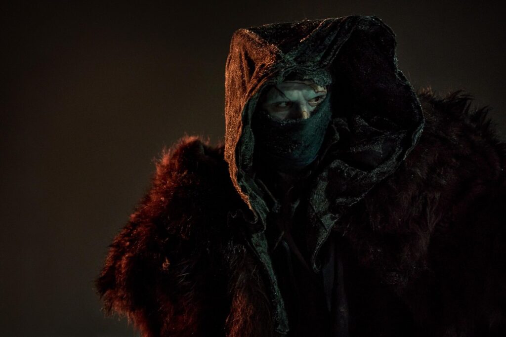 Cena do filme Frankenstein (2026), de Guillermo del Toro. O ator Jacob Elordi, interpretando o Monstro de Frankenstein, surge encapuzado, com o rosto parcialmente coberto por máscara e envolto em uma capa escura com detalhes de pele. A iluminação sombria reforça o ar enigmático e ameaçador da criatura.