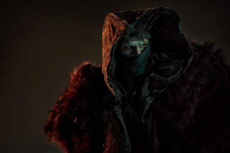 Cena do filme Frankenstein (2026), de Guillermo del Toro. O ator Jacob Elordi, interpretando o Monstro de Frankenstein, surge encapuzado, com o rosto parcialmente coberto por máscara e envolto em uma capa escura com detalhes de pele. A iluminação sombria reforça o ar enigmático e ameaçador da criatura.
