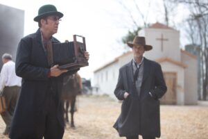 8 filmes de faroeste modernos em 2025: os lançamentos que renovam o gênero Nicolas Cage como Ben e Stephen Dorff como Thomas Keller em cena do filme Gunslingers (2025), um dos filmes de faroeste modernos em 2025. Eles estão em um vilarejo do Velho Oeste, vestidos com trajes típicos de pistoleiros — chapéus de aba larga, casacos escuros e botas de couro. Cage, à esquerda, segura uma antiga câmera fotográfica de madeira, enquanto Dorff observa à direita, diante de uma pequena igreja branca com cruz no topo e um cavalo parcialmente visível ao fundo. A iluminação quente e o cenário árido reforçam o clima clássico dos faroestes, reinterpretado sob uma estética contemporânea.