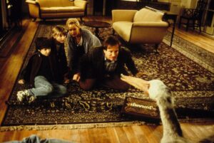 Cena do filme Jumanji (1995) mostrando Alan Parrish (Robin Williams) ajoelhado no tapete junto a Sarah Whittle (Bonnie Hunt), Judy Shepherd (Kirsten Dunst) e Peter Shepherd (Bradley Pierce) observando uma ave exótica que surge do tabuleiro mágico de Jumanji, dentro de uma sala iluminada e repleta de móveis de época.