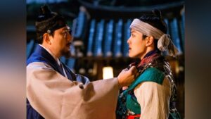 Cena intensa do dorama coreano de época famoso The Crowned Clown (2019), mostrando o ator Kim Sang-kyung, no papel de Yi Kyu, agarrando pela gola o personagem Ha-seon, interpretado por Yeo Jin-goo. Os dois homens, vestidos com trajes tradicionais da dinastia Joseon, estão frente a frente em um pátio iluminado por tochas, com a arquitetura coreana antiga visível ao fundo. A expressão de tensão e confronto entre mestre e súdito reflete o tom dramático característico dos doramas coreanos de época famosos, mesclando emoção, poder e lealdade.