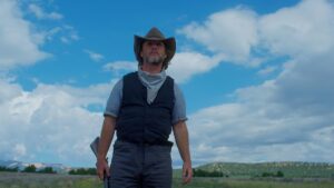 Imagem promocional do filme This Bloody Country (2024): ao centro, o personagem interpretado por Tim Blake Nelson aparece em pé sob um céu azul com nuvens densas, vestindo chapéu de caubói, colete escuro e lenço no pescoço, com uma espingarda apoiada ao lado do corpo — a paisagem árida e o horizonte distante reforçam a atmosfera clássica dos filmes de faroeste novos, combinando estética tradicional e drama contemporâneo do gênero.