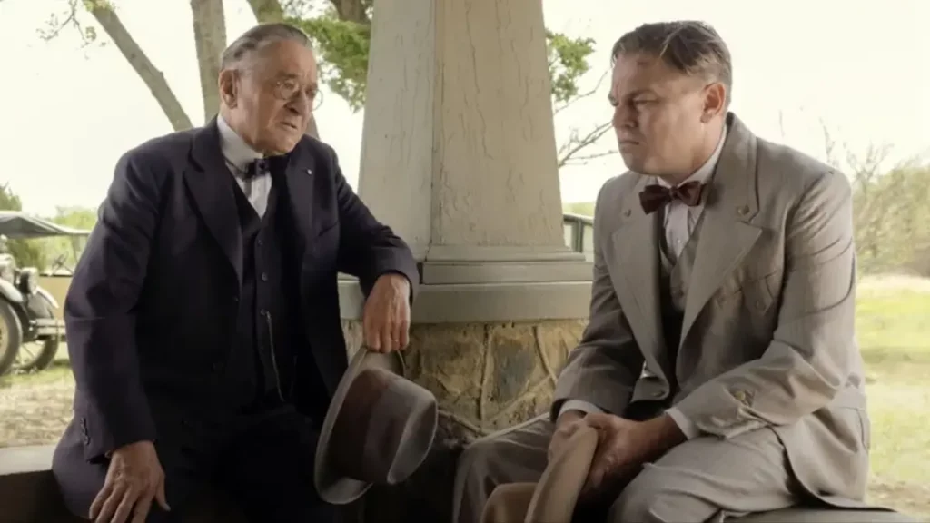 Cena do filme Assassinos da Lua das Flores: dois homens de terno sentados na varanda de uma casa conversam com expressões sérias, segurando chapéus, com árvores e um automóvel antigo ao fundo.
