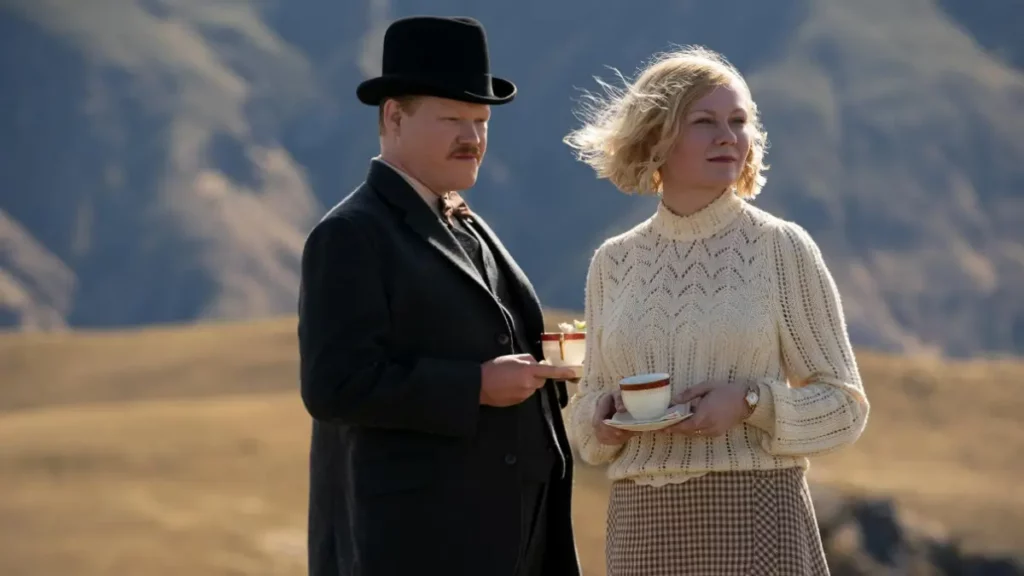 Cena do filme Ataque dos Cães: um homem de terno escuro e chapéu preto e uma mulher de suéter bege seguram xícaras de chá enquanto observam a paisagem montanhosa ao fundo.