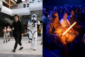 Comic-Con em Málaga reúne estrelas e momentos marcantes Registro da Comic Con Málaga 2025 com foco em cosplayers e público. À esquerda, participantes vestidos como personagens de Star Wars caminham pela área externa moderna da convenção, incluindo um stormtrooper com armadura branca e um fã caracterizado como Jedi. Ao fundo, outros visitantes observam e registram fotos. À direita, no interior de um auditório iluminado por luz azul, um cosplayer de Chewbacca segura um sabre de luz laranja aceso, cercado por espectadores atentos e sorridentes, reforçando o clima festivo e imersivo do evento.