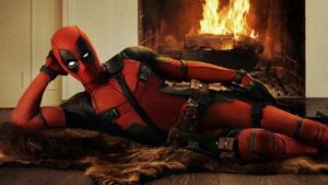 Deadpool deitado em frente a uma lareira, em uma pose descontraída, com o traje vermelho e preto característico.
