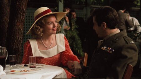 Diane Keaton como Kay Adams, vestindo um chapéu de palha com fita vermelha e um vestido vermelho de bolinhas brancas, sentada a uma mesa ao ar livre ao lado de Michael Corleone (Al Pacino) em uma cena de O Poderoso Chefão (1972).