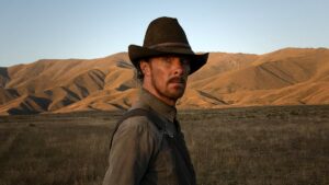 Assistir filme de faroeste: onde encontrar os melhores títulos online e na TV Cena do filme Ataque dos Cães (The Power of the Dog), perfeita para quem gosta de assistir filme de faroeste, mostrando o personagem Phil Burbank (interpretado por Benedict Cumberbatch) em um campo árido ao entardecer, usando chapéu de caubói e camisa marrom, com montanhas douradas ao fundo iluminadas pela luz suave do pôr do sol.