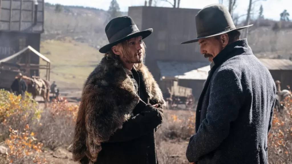 Cena de faroeste do filme Horizon: Uma Saga Americana - Capítulo 1. Dois homens conversam frente a frente ao ar livre. À esquerda, um homem de cabelos loiros compridos usa chapéu preto e um volumoso casaco rústico de pele. À direita, um homem mais velho com bigode veste chapéu cinza e um sobretudo grosso escuro. Ao fundo, há construções simples de madeira, carroças e vegetação seca típicas de um assentamento do velho oeste.