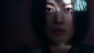 Close-up da protagonista de A Lição (2023), com expressão séria e iluminação parcial que reforça o clima sombrio da série.