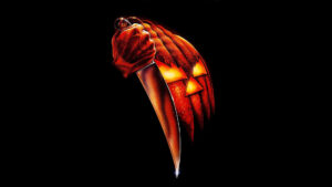 Pôster original de Halloween: A Noite do Terror, com uma abóbora e uma mão segurando uma faca sobre fundo preto.