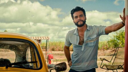 Wagner Moura em cena do filme O Agente Secreto, de Kleber Mendonça Filho, encostado em um carro amarelo em ambiente rural