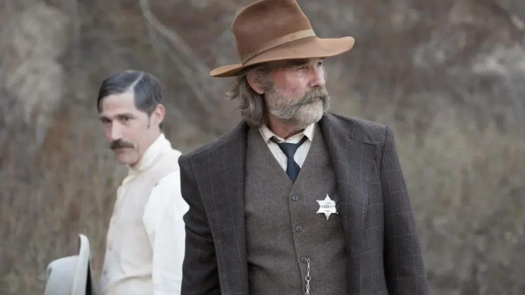 Captura de cena do filme Rastro de Maldade. Em primeiro plano à direita, o Xerife Franklin Hunt (interpretado por Kurt Russell), de barba branca e chapéu de cowboy marrom, terno e colete de tweed com uma estrela de xerife prateada, olha para a direita. No fundo desfocado à esquerda, Arthur O'Dwyer (interpretado por Patrick Wilson) está vestindo uma camisa branca e colete bege, segurando um chapéu de cowboy claro. O cenário é um terreno árido e rochoso com arbustos secos.