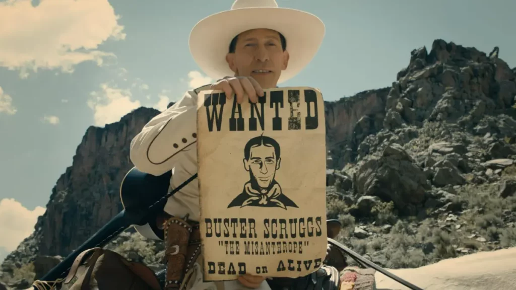 Close de Tim Blake Nelson como Buster Scruggs no filme The Ballad of Buster Scruggs. Ele segura um cartaz de procurado envelhecido com a inscrição "WANTED BUSTER SCRUGGS 'THE MISANTHROPE' DEAD OR ALIVE" e uma ilustração de seu rosto. Ele usa um chapéu de cowboy branco contra um fundo de montanhas rochosas e céu nublado.