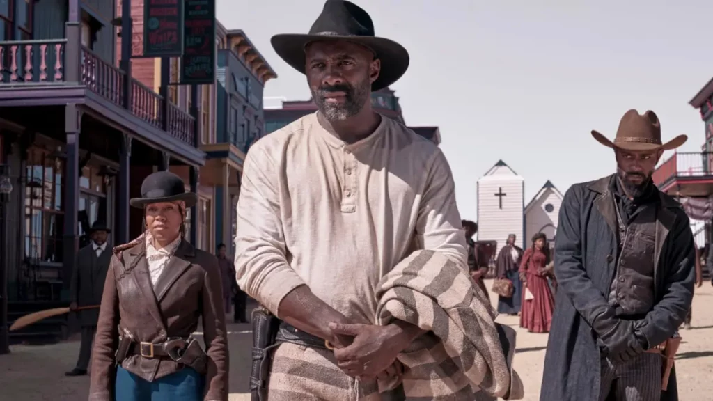 Cena de faroeste do filme Vingança & Castigo. Três personagens negros estão parados lado a lado em uma rua de terra. No centro, Idris Elba usa chapéu preto e camisa clara. À esquerda, Regina King veste chapéu-coco e jaqueta marrom. À direita, LaKeith Stanfield usa sobretudo preto e chapéu marrom. Ao fundo, vê-se construções de madeira típicas do velho oeste e uma pequena igreja.
