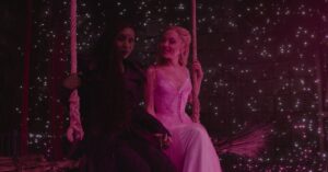 Elphaba e Glinda em cena de Wicked Parte 2 sentadas em um balanço sob luz rosa em ambiente estrelado.