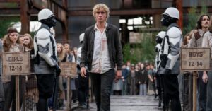 Jovem Haymitch caminha durante a seleção dos tributos em cena de Jogos Vorazes: O Amanhecer da Colheita, cercado por guardas e habitantes do distrito