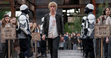 Jovem Haymitch caminha durante a seleção dos tributos em cena de Jogos Vorazes: O Amanhecer da Colheita, cercado por guardas e habitantes do distrito
