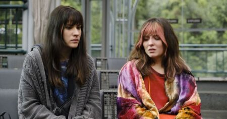 Duas jovens sentadas lado a lado em uma área externa, com expressões sérias, em cena do filme Traição Entre Amigas.