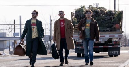 Kevin, Nick e Joe Jonas caminham lado a lado em cena do filme Um Natal Bem Jonas Brothers, carregando malas enquanto passam por uma área externa.