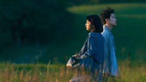 Um homem e uma mulher observam o horizonte ao entardecer, cercados por um campo verde.