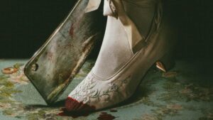close-up de um sapato branco de cetim com bordado floral, coberto por sangue na ponta, ao lado de um cutelo manchado de sangue sobre um tapete verde