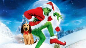 personagem verde com roupa de Papai Noel segura uma bola natalina ao lado de um cachorro com laço vermelho em cenário nevado.