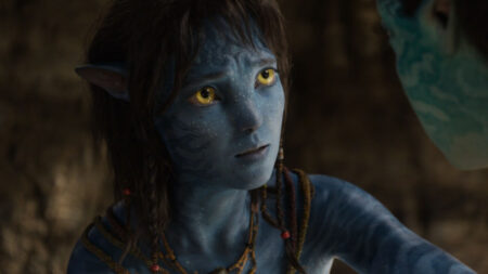 Avatar: Personagem Na’vi de pele azul e olhos amarelos olha com expressão atenta em ambiente de pouca luz.