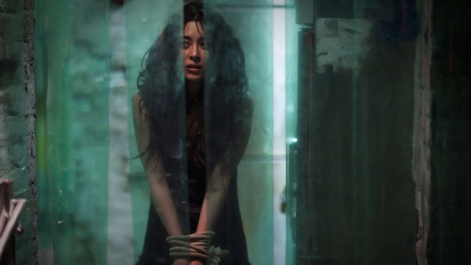 Filmes de terror asiáticos famosos: 12 obras que conquistaram o mundo mulher com as mãos amarradas em um ambiente escuro e úmido, observada por trás de cortinas translúcidas verdes, em cena de tensão psicológica.