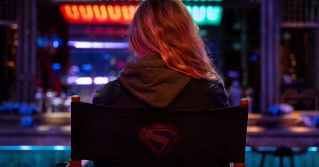 Milly Alcock de costas sentada em cadeira de diretor com o símbolo da Supergirl no set iluminado.