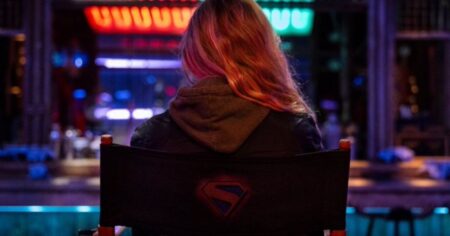 Milly Alcock de costas sentada em cadeira de diretor com o símbolo da Supergirl no set iluminado.