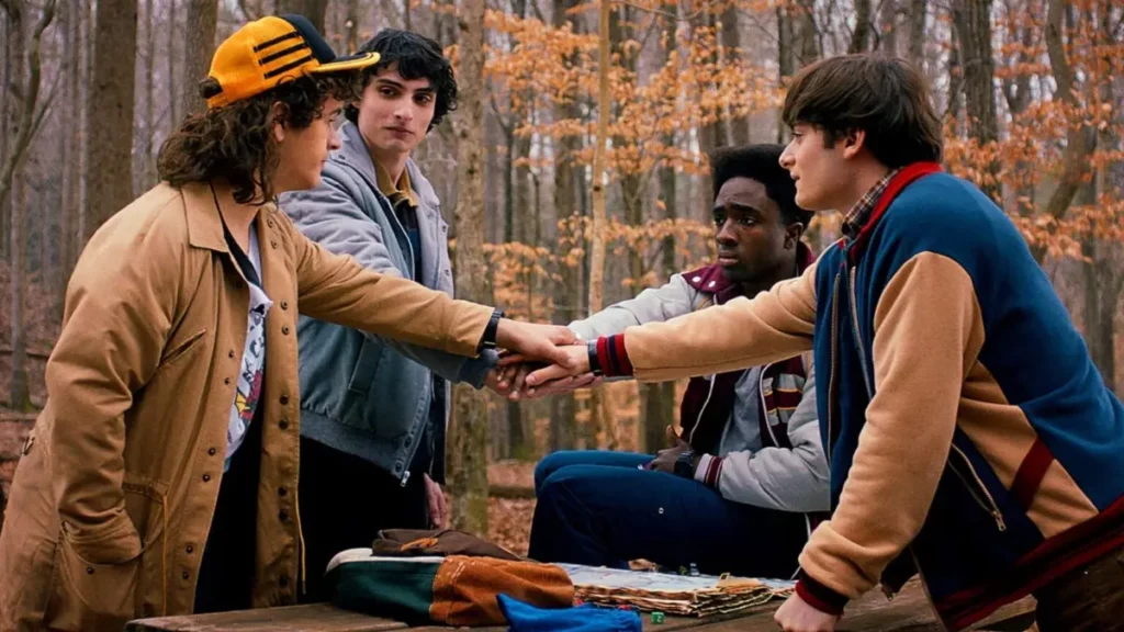 Mike, Dustin, Lucas e Will unem as mãos durante uma cena de Stranger Things em uma área de floresta em Hawkins.