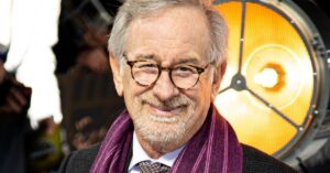 Spielberg admite arrependimento ligado a filme ganhador do Oscar; entenda Steven Spielberg sorri ao posar para fotos em evento público, usando óculos de armação redonda, cachecol roxo e terno escuro, com iluminação de estúdio ao fundo