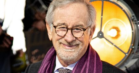 Spielberg admite arrependimento ligado a filme ganhador do Oscar; entenda Steven Spielberg sorri ao posar para fotos em evento público, usando óculos de armação redonda, cachecol roxo e terno escuro, com iluminação de estúdio ao fundo