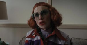 Amy Madigan surpreende no Critics Choice e vence Ariana Grande; conheça a atriz Amy Madigan caracterizada como tia Gladys em cena do filme A Hora do Mal, usando óculos grandes e cabelo ruivo, sentada em um ambiente interno com expressão séria