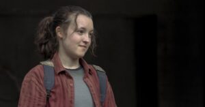 Bella Ramsey caracterizada com mochila e camisa vermelha em cena dramática, com expressão séria e iluminação escura ao fundo