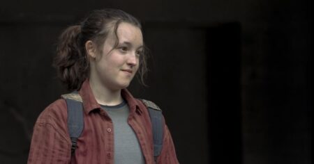 Bella Ramsey comenta desejo de viver Homem-Aranha no cinema; veja Bella Ramsey caracterizada com mochila e camisa vermelha em cena dramática, com expressão séria e iluminação escura ao fundo